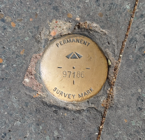 Close-up view of a small, round, dull brass-coloured metal disc with engraved words 'permanent survey mark' and the number 97186 visible, embedded in concrete.