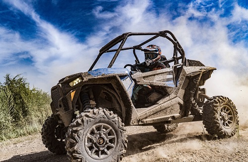 Person wearing a helmet and protective clothing dirving a mud-covered side-by-side vehicle outdoors