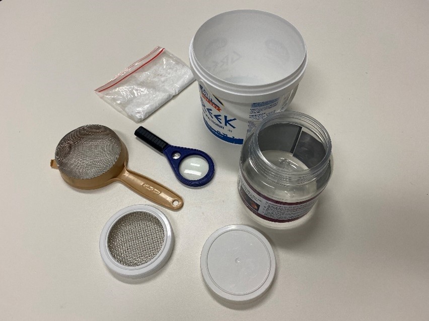 Photo of equipment used to create a sugar shake kit. Equipment includes shake jar, mesh and solid lids, strainer, small bucket, pure icing sugar and magnifying glass.